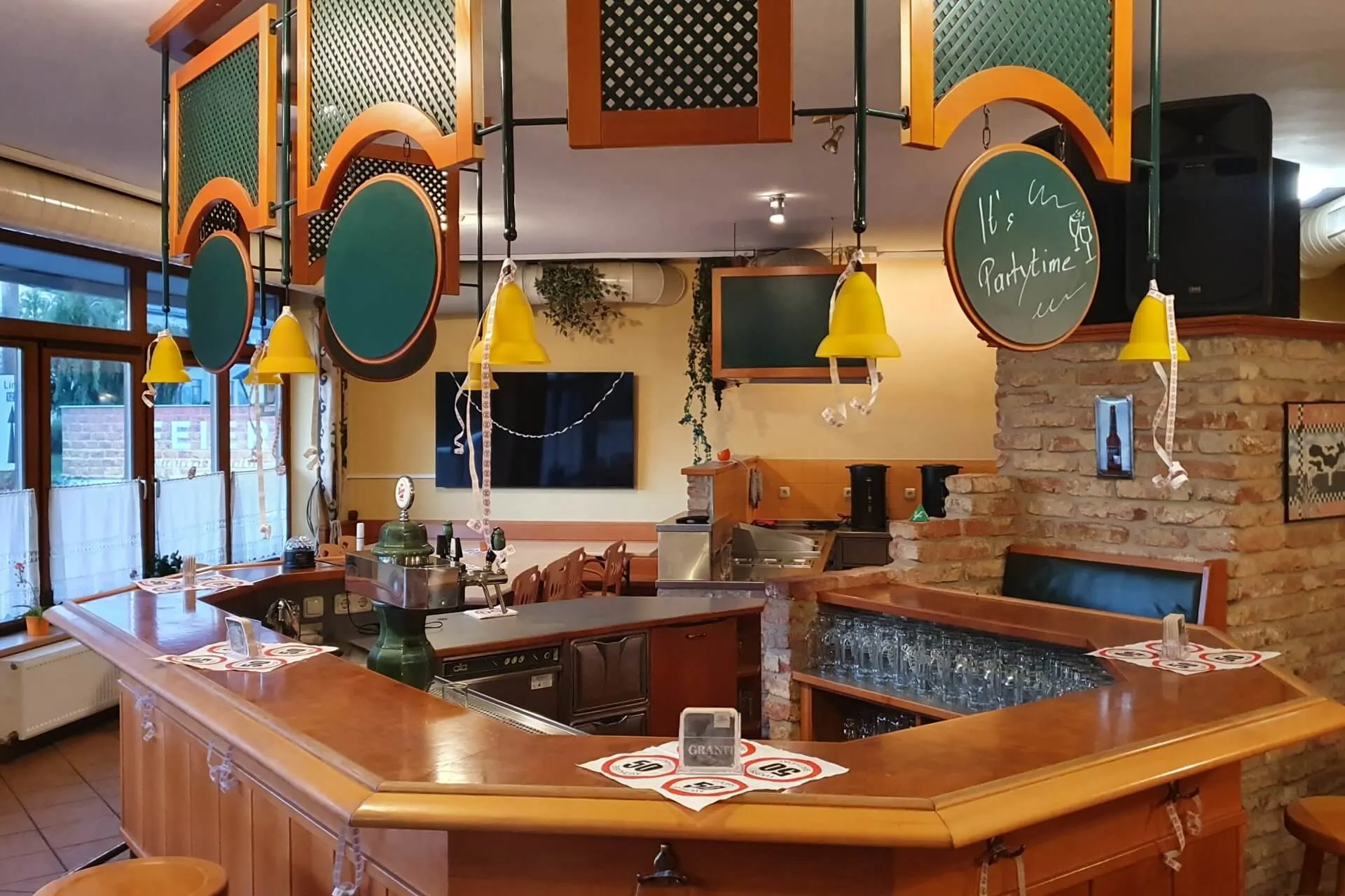 Eine gemütliche Bar mit einem hölzernen Tresen, gelben Hängelampen, runden grünen Schildern und Glaswaren in Regalen. Auf einer Kreidetafel steht "It's Parttime". Große Fenster lassen natürliches Licht herein.