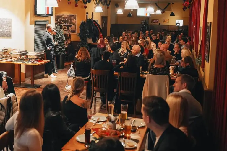 Eine lebhafte Restaurantszene zeigt Gäste, die an Tischen sitzen, essen und einen Redner beobachten, der vorne im Raum steht. Warme Beleuchtung und festliche Dekoration schaffen eine gemütliche, einladende Atmosphäre.
