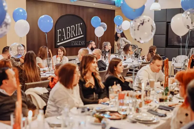 Eine Gruppe von Menschen sitzt an geschmückten Tischen mit Speisen und Getränken bei einer lebhaften Veranstaltung in einer Halle. An der Wand im Hintergrund hängen blaue und weiße Luftballons, und auf einem großen Schild steht FABRIK EKSKLUZIV.