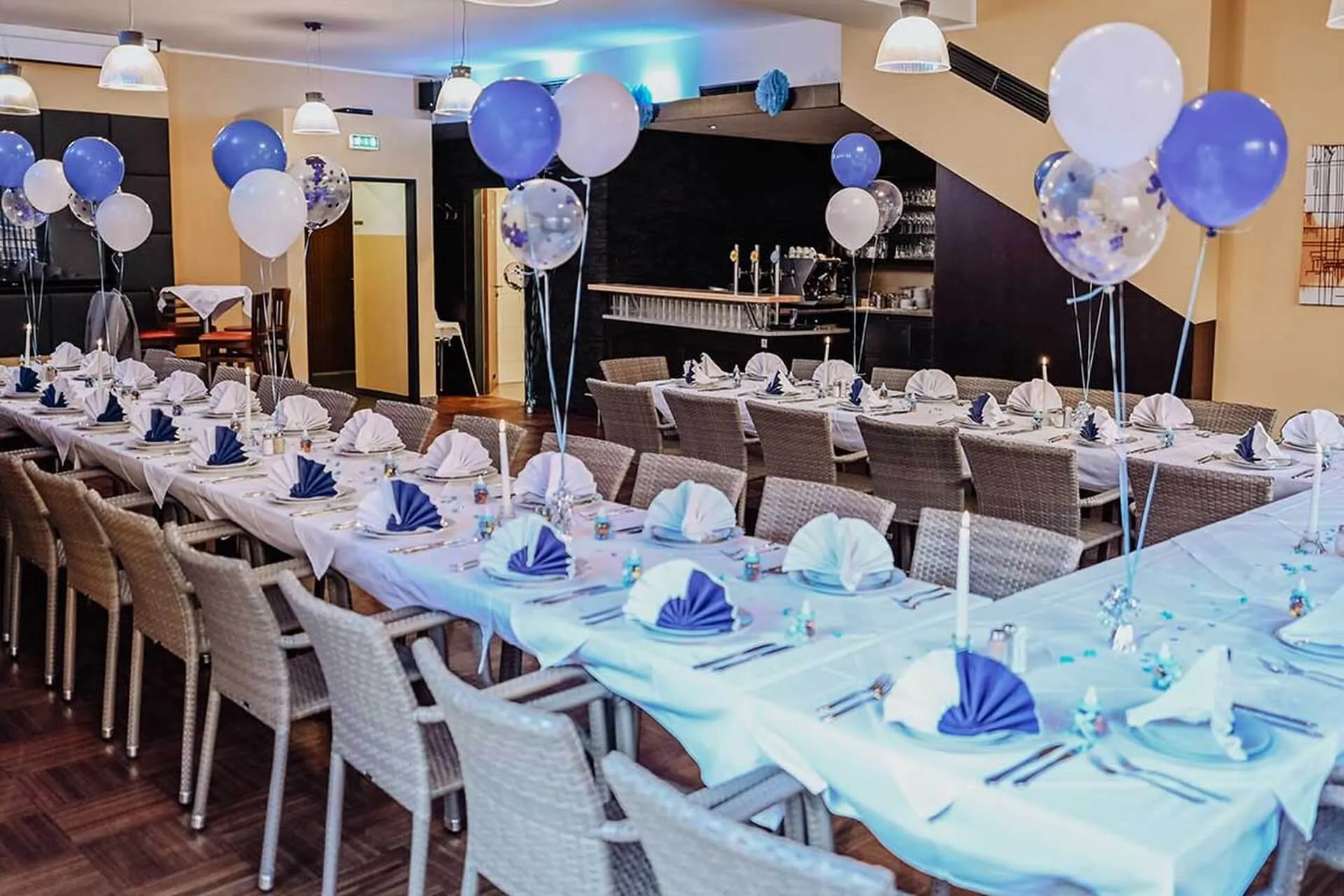 Zwei lange Banketttische sind für eine Party mit weißen Tischtüchern, blauen und weißen Servietten und Ballon-Mittelstücken in Blau, Weiß und Transparent gedeckt. Korbstühle umgeben die Tische in einem hell erleuchteten Raum.
