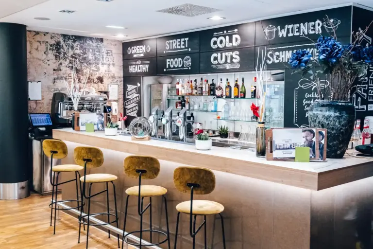 Eine moderne Bar mit vier gelben Hockern, einem hölzernen Tresen, Flaschen und Gläsern in Regalen, Wandmenüs im Kreidestil und dekorativen Vasen. Die Atmosphäre ist hell und einladend.
