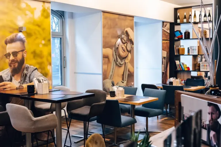Modernes Café-Interieur mit leeren Tischen und Stühlen, großen Wandpostern mit Menschen, Bücherregalen mit Flaschen und Büchern und natürlichem Licht, das durch ein Fenster fällt.