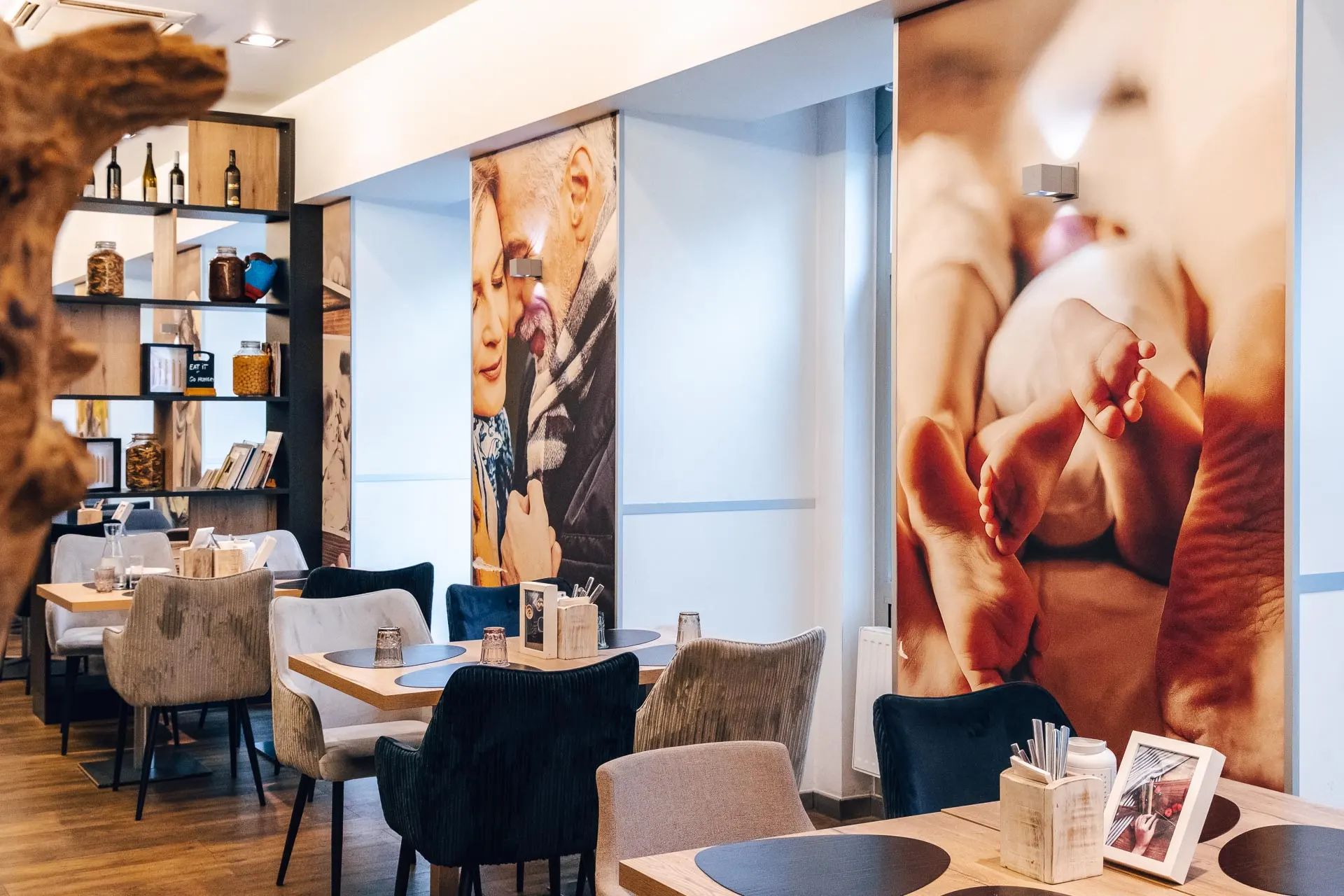 Ein modernes, gemütliches Restaurant mit gepolsterten Stühlen, Holztischen, Regalen mit Dekoration und Büchern sowie großen Wandbildern mit Nahaufnahmen von Menschen und Babyfüßen. Die warme Beleuchtung schafft eine einladende Atmosphäre.
