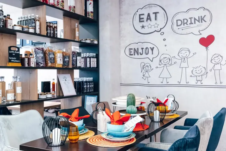 Ein gemütliches Café mit modernem Design, Regalen mit Lebensmitteln und einem mit bunten Servietten, gewebten Tischsets und kleinen Zierkakteen gedeckten Tisch. Ein Wandgemälde zeigt lächelnde Strichmännchen und die Worte Eat, Drink, and Enjoy.