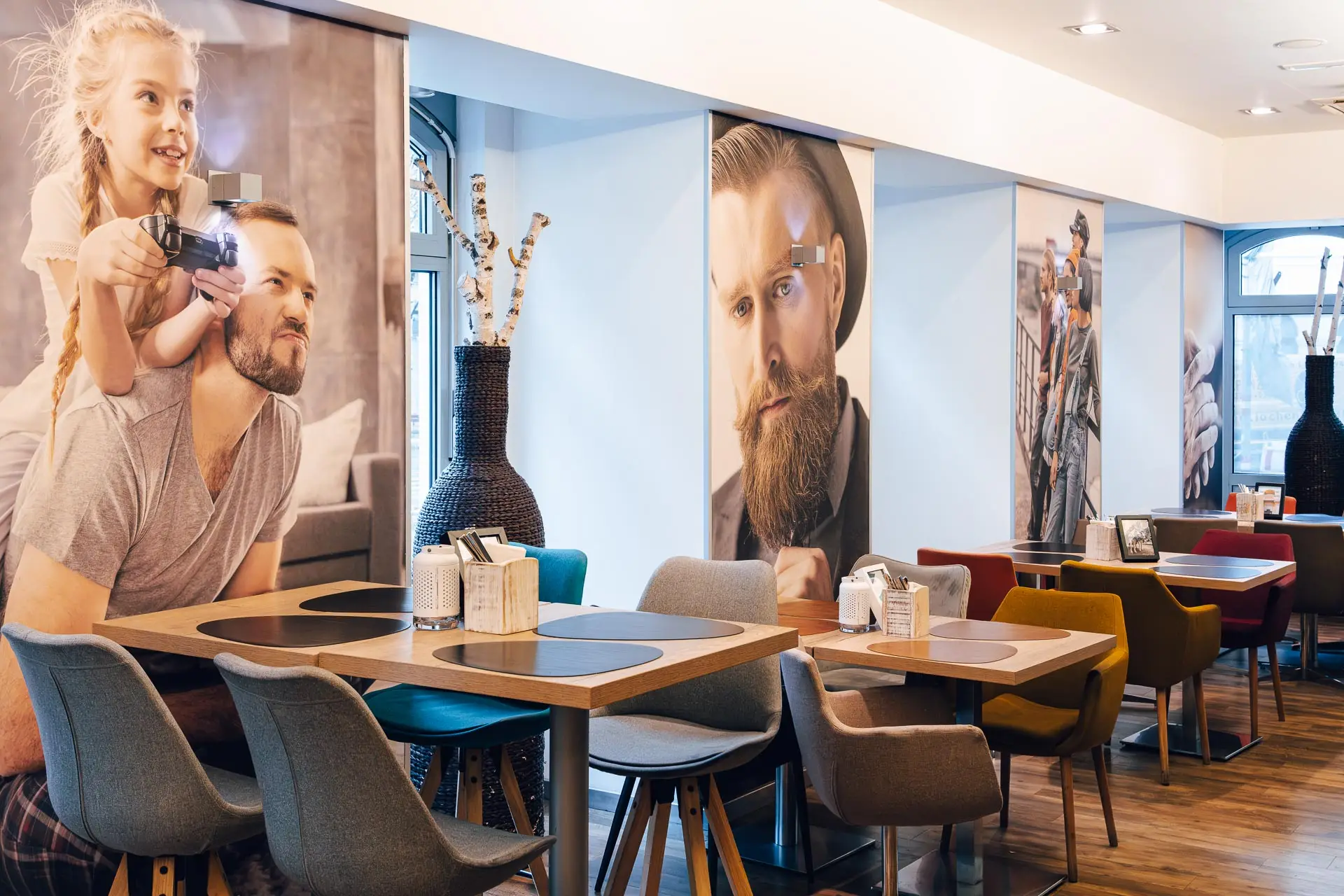 Ein modernes, hell beleuchtetes Café mit bunten Stühlen und Tischen. Große Wandgemälde zeigen Menschen, darunter einen Mann mit einem Kind auf dem Rücken und einen bärtigen Mann. Gedeckte Tische und dekorative Vasen tragen zur gemütlichen Atmosphäre bei.
