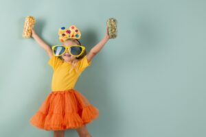 Ein junges Mädchen mit einer übergroßen Sonnenbrille, einem gelben Stirnband mit Schleife, einem gelben Hemd und einem orangefarbenen Tutu hebt die Arme mit Dosen von Popcorn, bereit zum Feiern mit ihrem Lieblings-Faschingsrezept vor einem hellblauen Hintergrund.