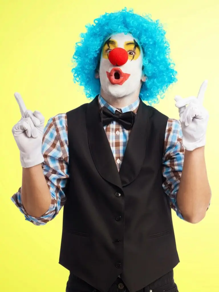Eine als Clown gekleidete Person mit blauer Lockenperücke, roter Nase, weißer Gesichtsbemalung und bunter Schminke trägt ein kariertes Hemd, eine schwarze Weste, eine Fliege und weiße Handschuhe und deutet mit beiden Zeigefingern auf eine Faschingsfeier vor einem gelben Hintergrund.