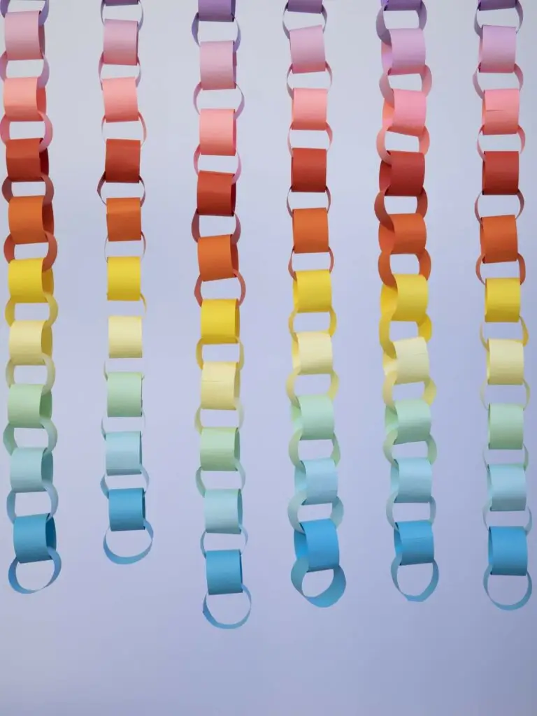 Sechs vertikale Papierketten aus pastellfarbenen Schleifen hängen vor einem hellvioletten Hintergrund. Perfekt für eine Faschingsfeier ist jede Kette mit einem Farbverlauf aus Rosa, Gelb, Grün, Blau und Orange versehen.