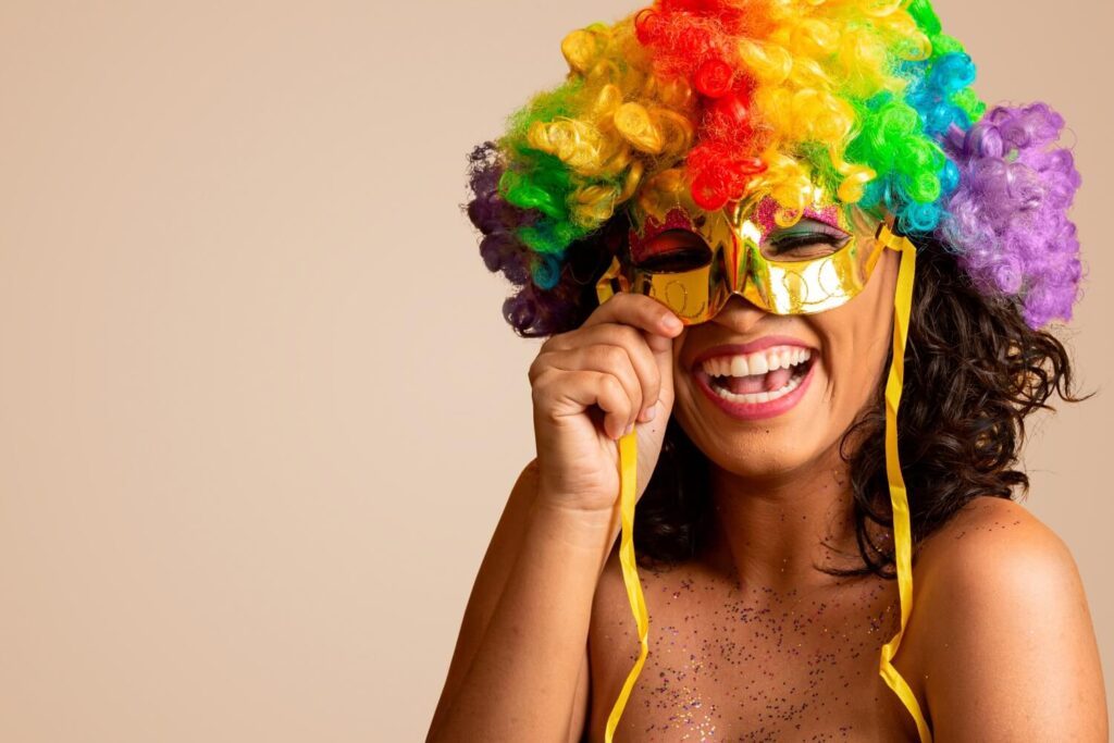 Eine Person, die eine bunte, gelockte Clownsperücke und eine goldglänzende Maske trägt, lächelt strahlend und hält das Band der Maske. Ihre glitzernde Haut und ihr festliches Aussehen erinnern an den Geist der Faschingsfeier, die vor einem schlichten hellbeigen Hintergrund stattfindet.