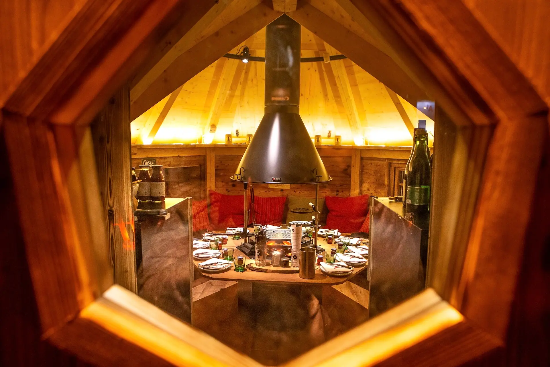 Der Blick durch ein sechseckiges Fenster auf das gemütliche Interieur einer Holzhütte: ein runder Tisch, der für eine Mahlzeit gedeckt ist, rote Kissen und ein großer Grill in der Mitte, alles in warmem Licht getaucht.