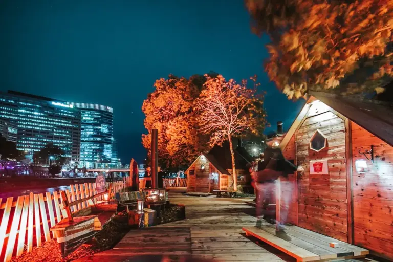Feuerdorf am Donaukanal Eine hell erleuchtete Flusslandschaft bei Nacht mit Holzhütten, herbstlich gefärbten Bäumen und einem modernen Glasgebäude im Hintergrund. Eine verschwommene Person geht einen Holzsteg entlang.