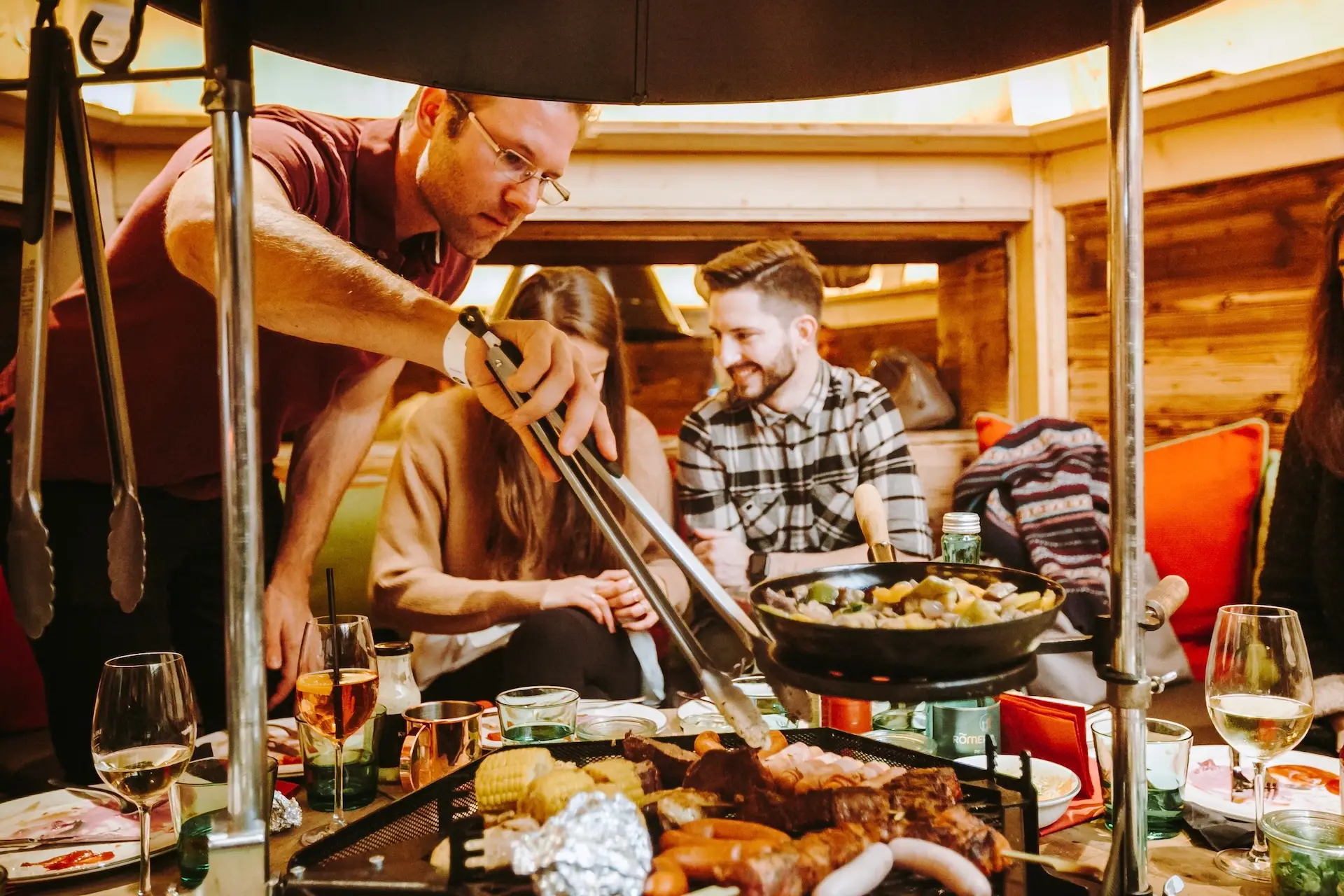 Ein Mann bereitet mit einer Zange Speisen auf einem Tischgrill zu, während zwei Personen lächelnd daneben sitzen. Auf dem Tisch stehen verschiedene Gerichte, Getränke und Besteck in einer gemütlichen Holzumgebung.