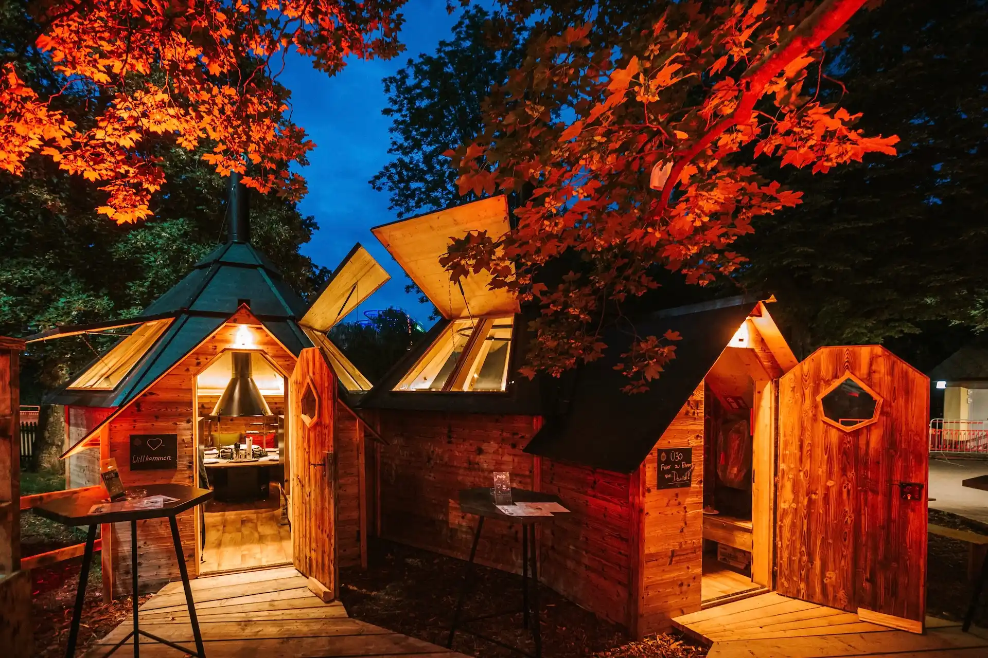 Drei kleine Holzhütten mit offenen Türen und warm leuchtendem Licht stehen in der Abenddämmerung unter Bäumen mit orangefarbenen Blättern und schaffen eine gemütliche, einladende Atmosphäre.