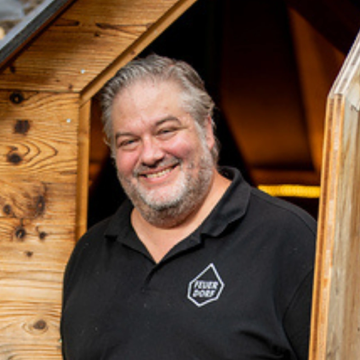 Ein lächelnder Mann mit grauen Haaren und Bart steht am Eingang eines Holzgebäudes und trägt ein schwarzes Hemd mit einem sechseckigen Feuerdorf-Logo darauf.