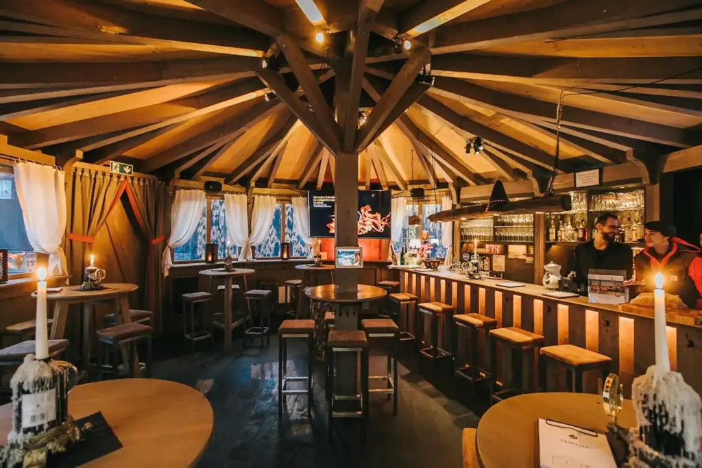 Gemütliche Holzbar mit runden Tischen, hohen Barhockern, einer Decke mit zentralen Holzbalken, warmer Beleuchtung und Menschen hinter der Theke. An den Fenstern hängen Vorhänge, die eine einladende Atmosphäre schaffen.