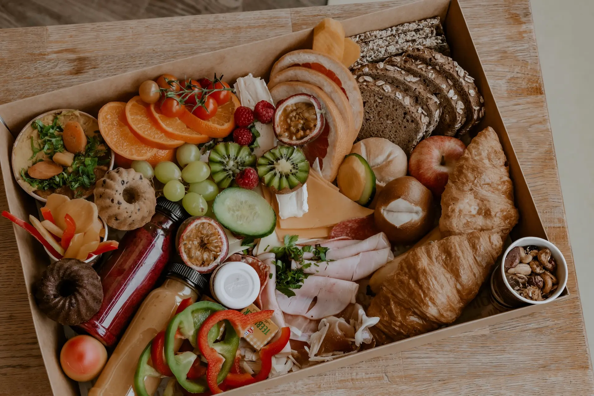 Eine Frühstücksbox mit Croissants, geschnittenem Brot, Käse, Wurstwaren, frischem Obst, Gemüse, Dips, Müsli und kleinen Kuchen, alles fein säuberlich auf einem Holztisch angeordnet.