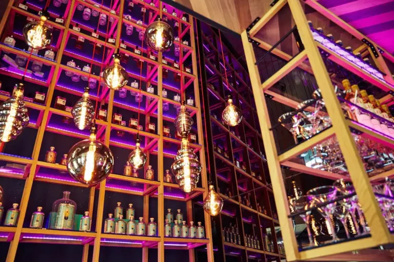 Eine stilvolle Bar mit Regalen voller Spirituosenflaschen, die von warmen Hängelampen und rosa-violetter Neonbeleuchtung beleuchtet werden und eine moderne und lebendige Atmosphäre schaffen. Die Gläser sind auf der rechten Seite zu sehen.
