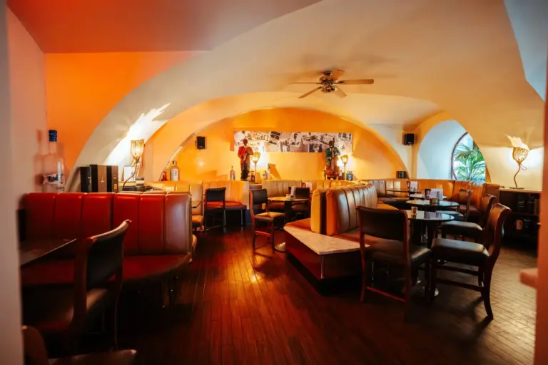 First American Bar Gemütliches Restaurantinterieur mit gewölbten Decken, warmer gelber Beleuchtung, Sitznischen mit roten und beigen Sitzgelegenheiten, Holzböden und einer Fotocollage an der Rückwand. Die Tische sind gedeckt, aber es sind keine Gäste im Raum.