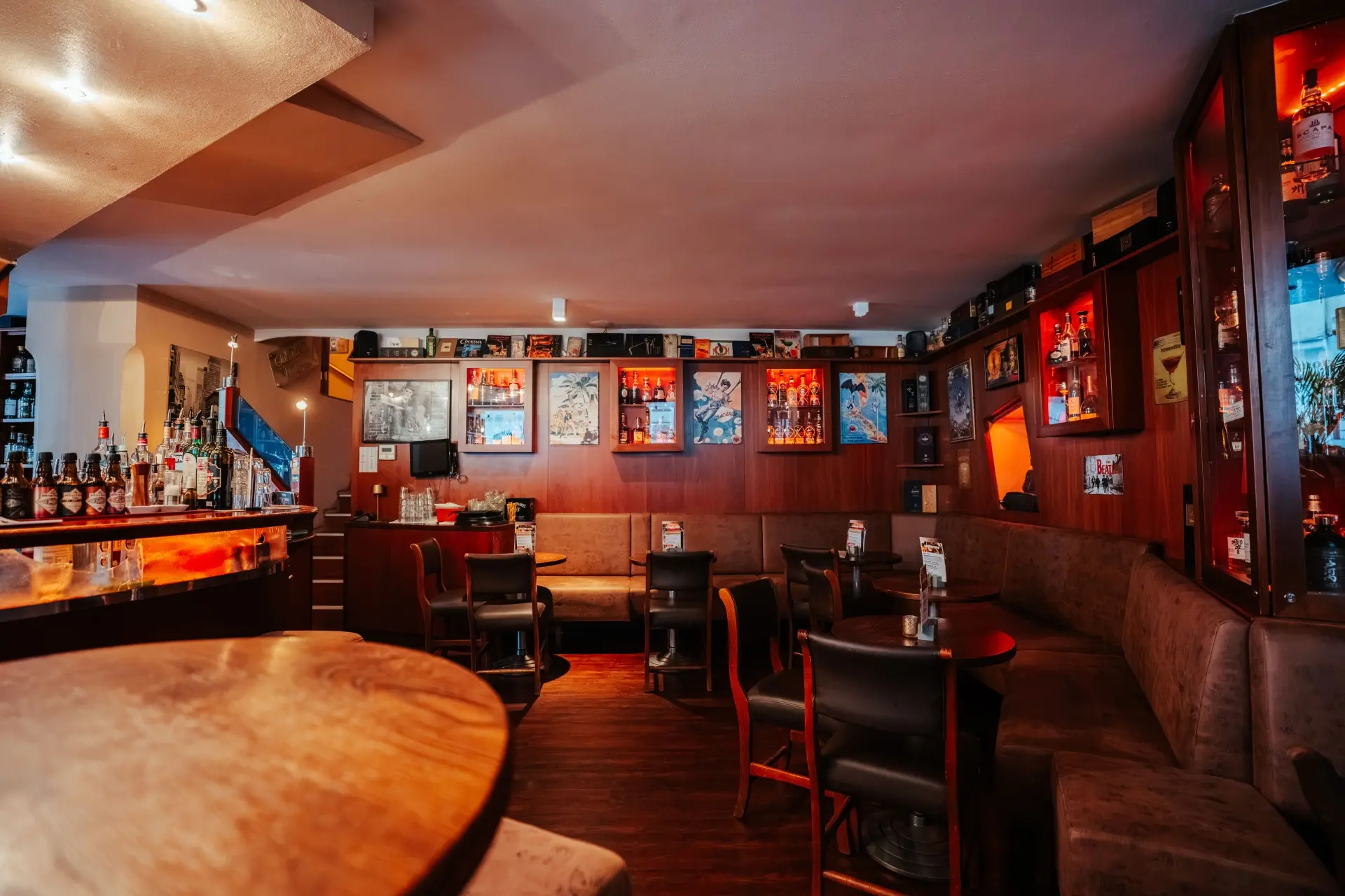 Gemütliches Bar-Interieur mit Holztischen, gepolsterten Bänken und Stühlen. An den Wänden reihen sich Regale mit Flaschen und gerahmten Fotos aneinander. Warme Beleuchtung sorgt für eine entspannte, einladende Atmosphäre.