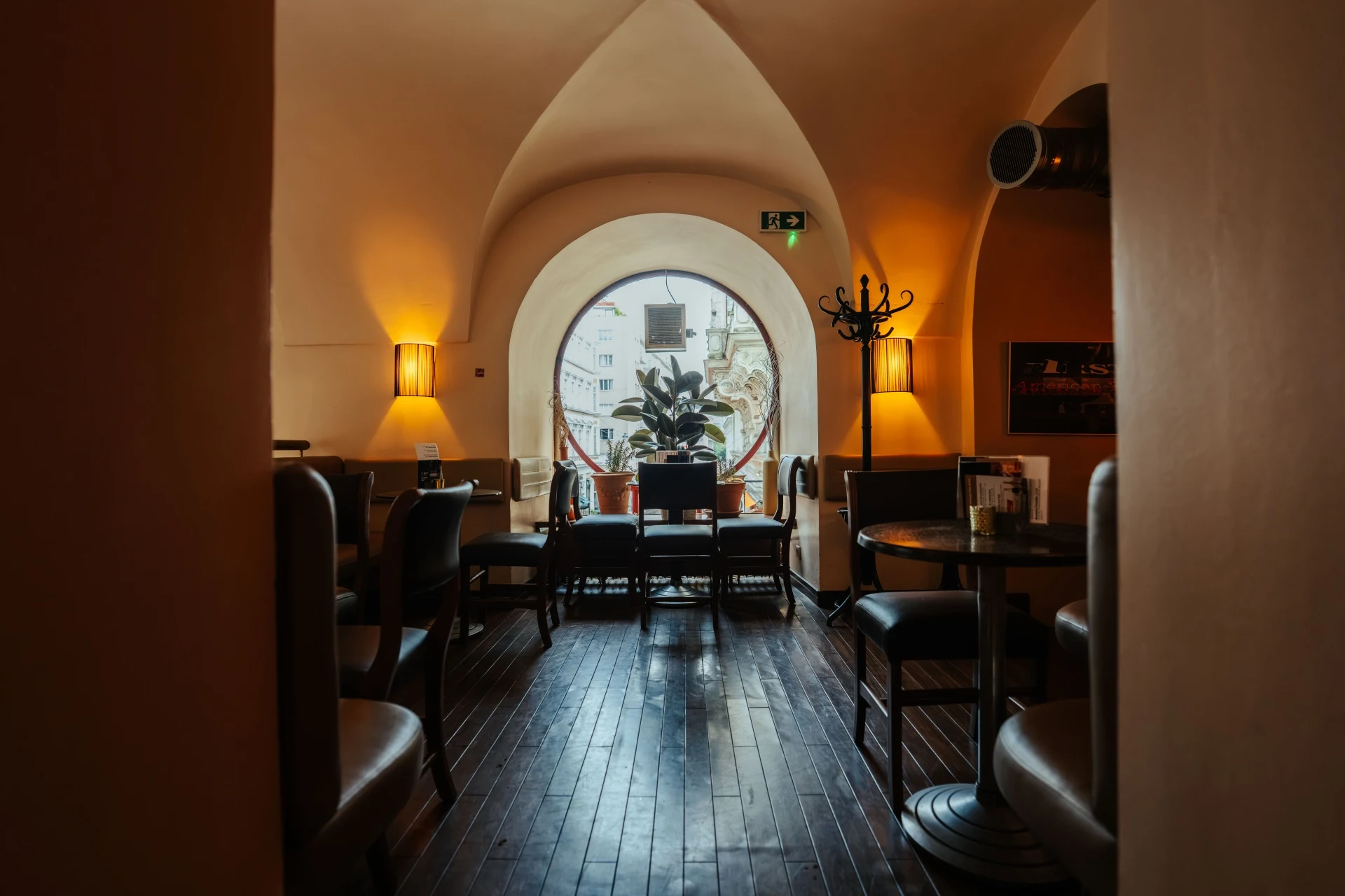 Gemütliches Café-Interieur mit dunklen Stühlen und Tischen, warmer Beleuchtung und einem großen runden Fenster im hinteren Bereich, durch das eine Topfpflanze und die Gebäude draußen zu sehen sind.