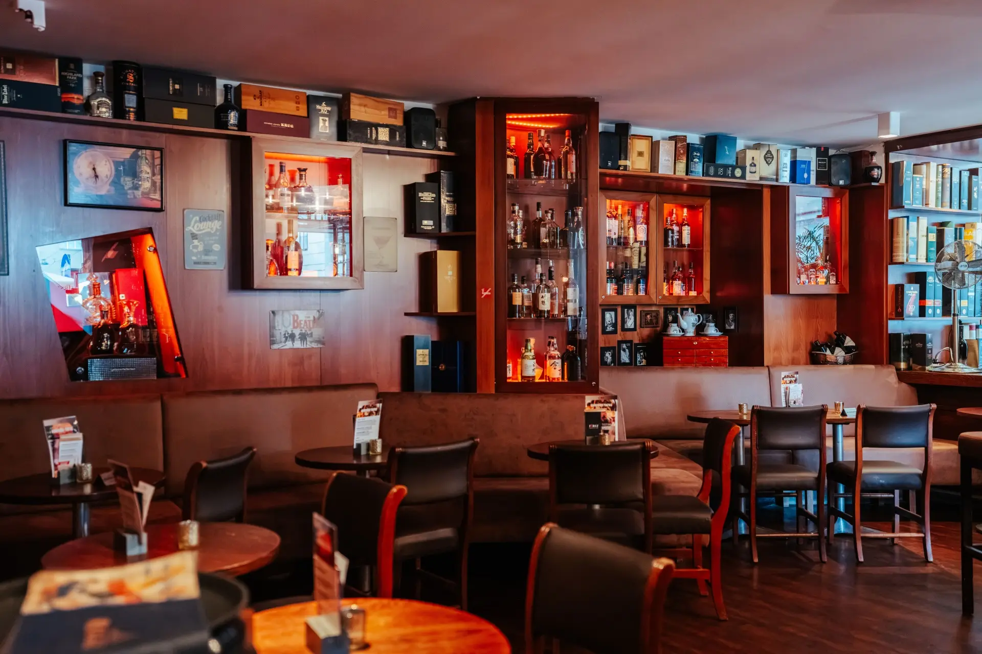 First American Bar Eine gemütliche Bar mit Holztischen und -stühlen, gedämpfter gelber Beleuchtung und Regalen hinter der Theke, die mit verschiedenen Spirituosenflaschen gefüllt sind. Die Atmosphäre ist warm und einladend, mit ordentlich angeordneten Sitzbereichen.