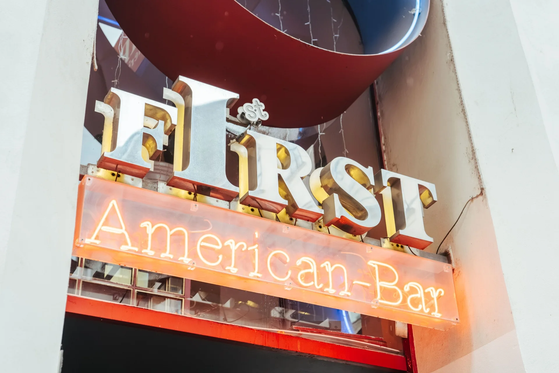 Ein Schild mit der Aufschrift „First American-Bar“ in Gold- und Neonbuchstaben, das über einem Eingang angebracht ist. Das Schild wird von einer weißen Wand und einer darüber liegenden, geschwungenen, rot-blauen Zierkonstruktion eingerahmt.