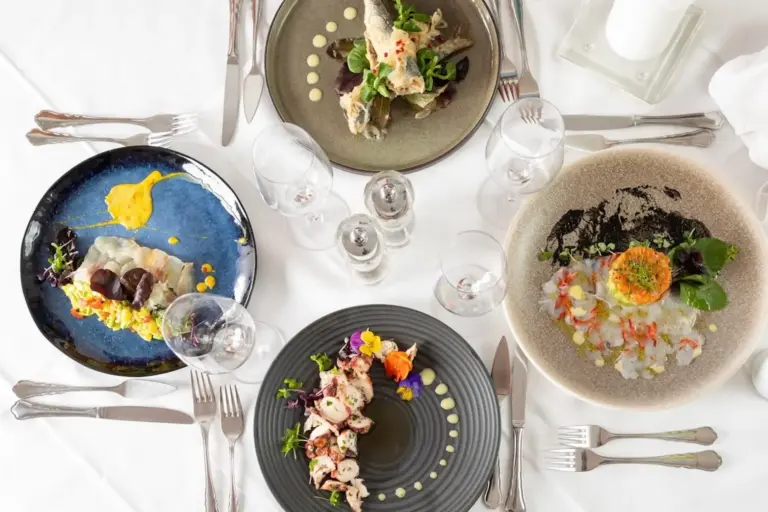 Drei elegante Teller mit Gourmetgerichten sind auf einem weißen Tischtuch angerichtet, umgeben von Besteck, Weingläsern sowie Salz- und Pfefferstreuern, so dass ein edles Ambiente entsteht.