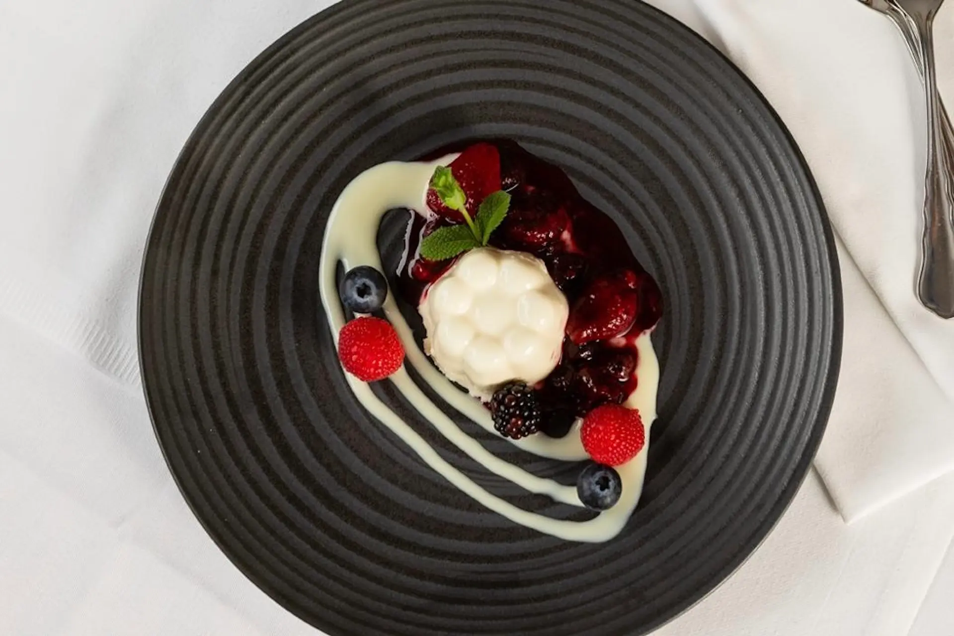 Ein schwarzer Teller mit einer weißen Panna Cotta in der Mitte, belegt mit gemischter Beerensauce, frischer Minze, Blaubeeren und Himbeeren und beträufelt mit weißer Sauce, auf einem weißen Tischtuch.