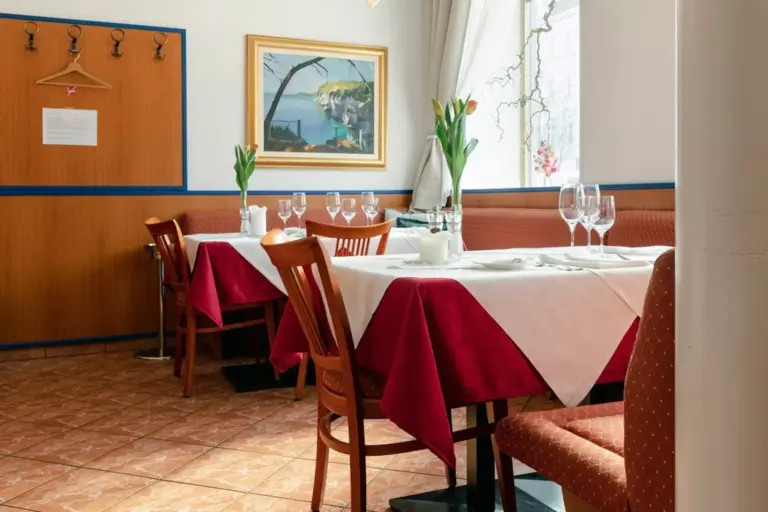 Ein gemütliches Restaurant mit weiß und rot gedeckten Tischen, Holzstühlen, Weingläsern, Vasen mit Tulpen und Kunstwerken an der Wand. Sonnenlicht strömt durch ein Fenster mit durchsichtigen Vorhängen herein.