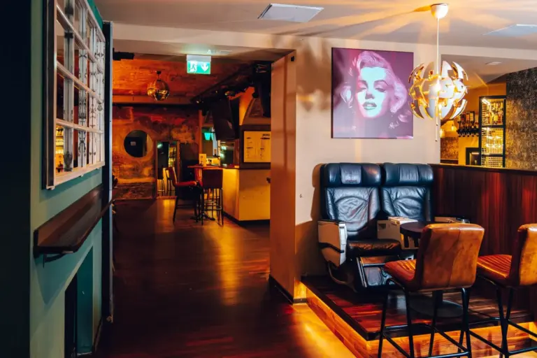 Eine gemütliche Bar mit Ledersesseln, Holzfußboden, gedämpfter Beleuchtung und einem Pop-Art-Porträt an der Wand. Es gibt Tische, Stühle und eine einzigartige Hängelampe, während im Hintergrund der Barbereich zu sehen ist.