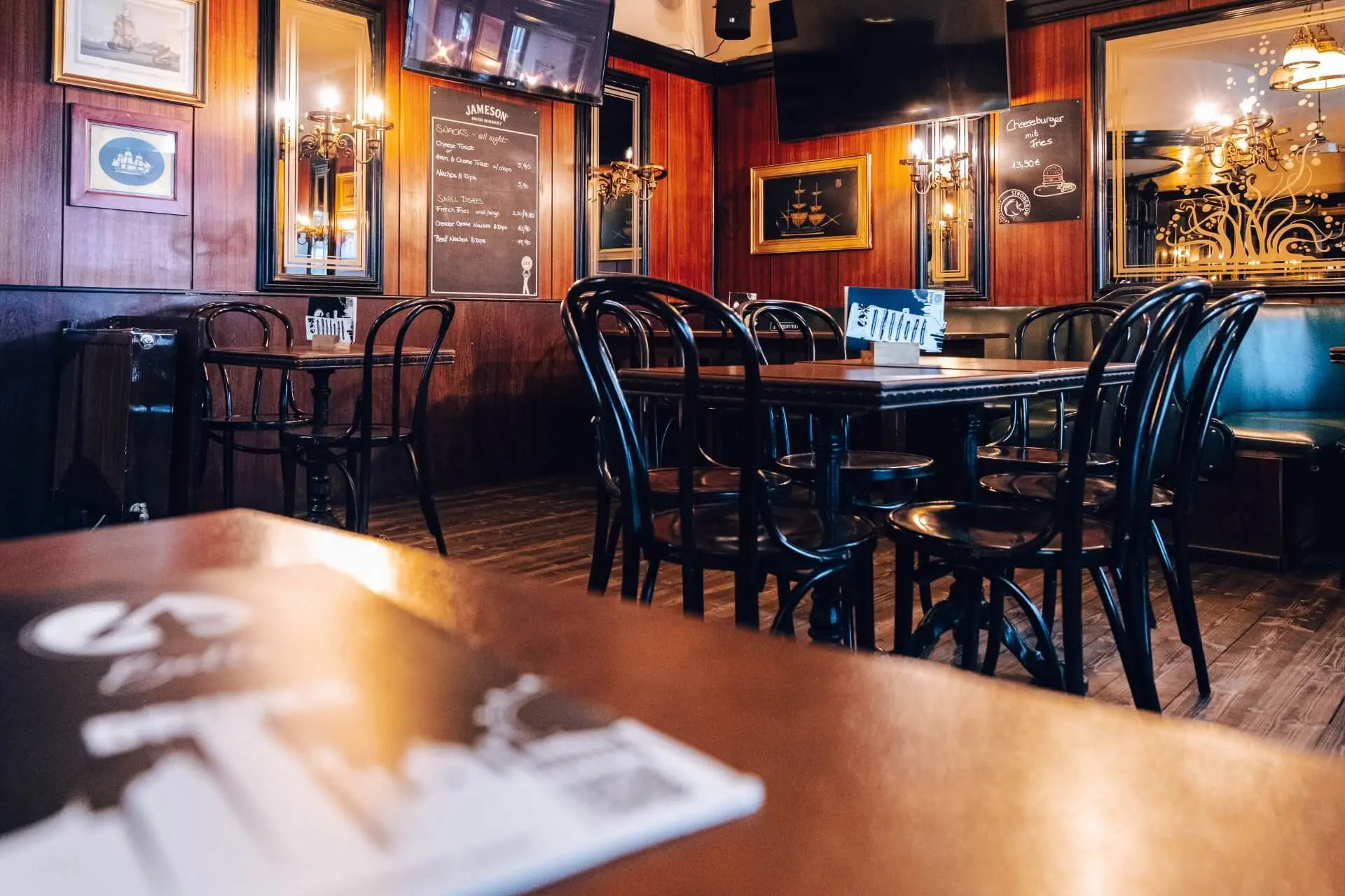 Leeres, gemütliches Restaurant mit Holzwänden und dunklen Holzstühlen und -tischen. Weiches, warmes Licht, gerahmte Bilder, große Spiegel und eine Speisekarte im Vordergrund schaffen eine einladende Atmosphäre.