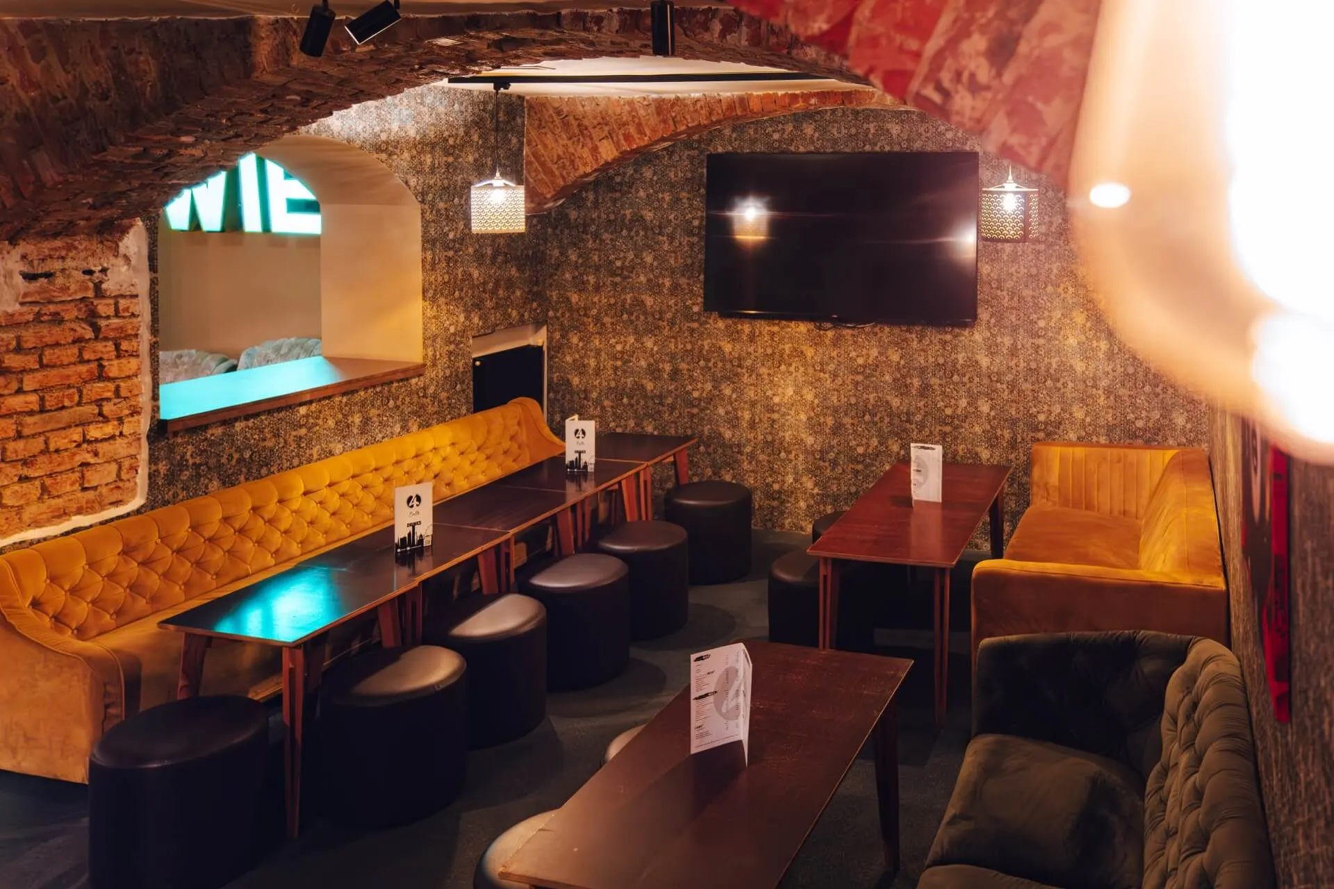 Eine gemütliche Lounge mit freiliegenden Backsteinbögen, Samtsofas, dunklen Holztischen, schwarzen runden Hockern, einem großen, an der Wand montierten Fernseher und warmer Beleuchtung, die eine einladende Atmosphäre schafft.