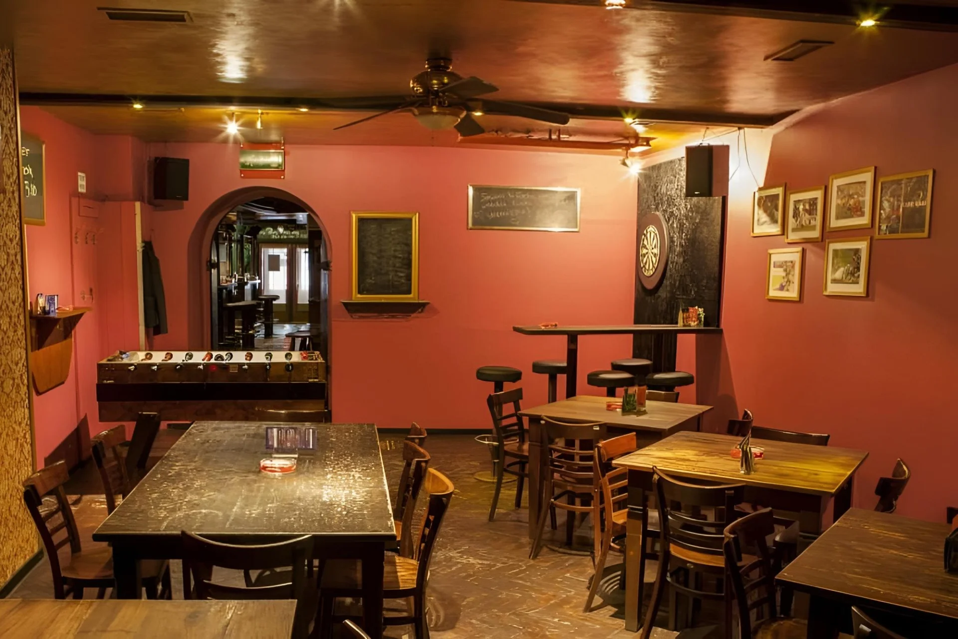 Eine gemütliche Bar mit rosafarbenen Wänden, Holztischen und -stühlen, einem Kickertisch, einer Dartscheibe, gerahmten Bildern, Kreidetafeln und warmer Deckenbeleuchtung schafft eine entspannte Atmosphäre.