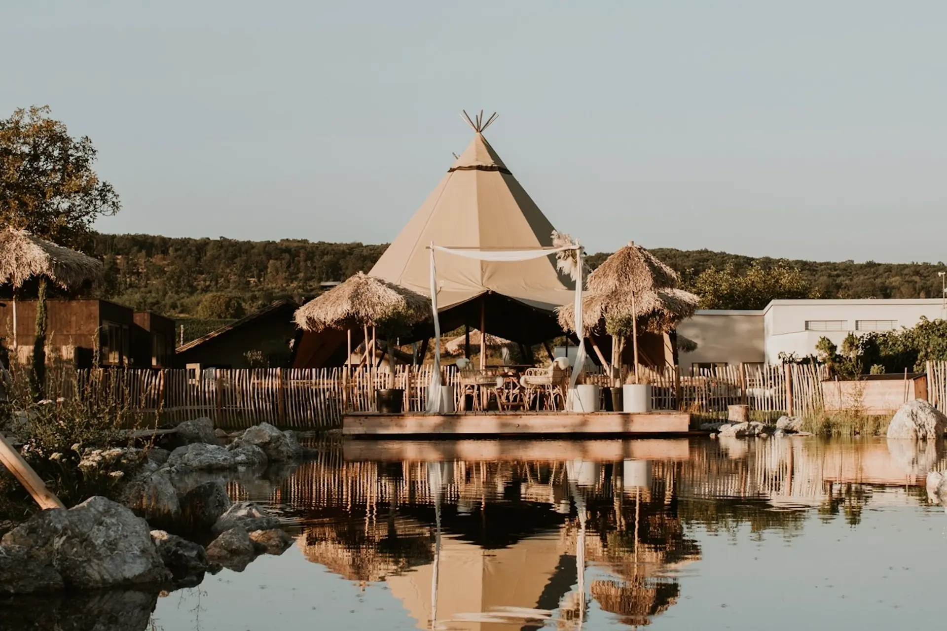 Franz von Grün Ein großes beigefarbenes Tipi-Zelt mit offenem Vordach und strohgedeckten Schirmen steht an einem ruhigen See, in dem sich die Struktur im Wasser spiegelt, umgeben von Felsen und Grünpflanzen unter einem klaren Himmel.