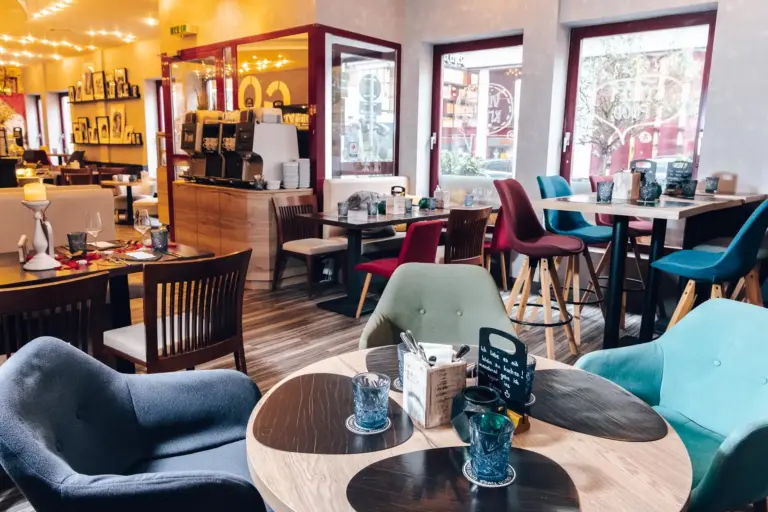 Ein gemütliches, modernes Café mit bunten Stühlen, runden Holztischen und großen Fenstern, die natürliches Licht hereinlassen. Im Hintergrund sind Kaffeemaschinen und Regale mit Bechern zu sehen.