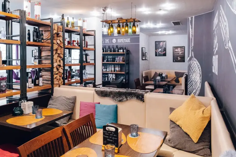 Ein gemütliches Restaurant mit gepolsterten Stühlen, Holztischen, farbenfrohen Kissen, Regalen mit Wein und Brennholz, modernen Hängelampen und Wanddekoration im Stil von Kreidetafeln. Die Atmosphäre ist warm und einladend.