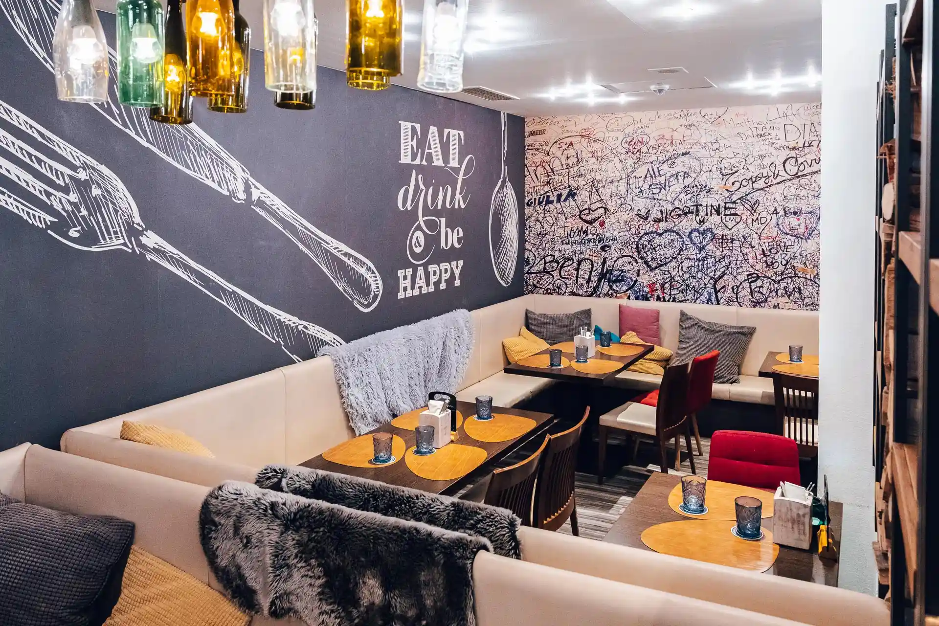 Ein gemütliches Restaurant mit gepolsterten Bänken, Holztischen und stimmungsvollem Licht aus hängenden Flaschen. Auf einer Kreidetafel steht EAT, drink & be HAPPY; eine andere Wand ist mit handgeschriebenen Botschaften und Kritzeleien bedeckt.