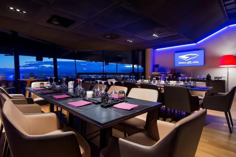 Ein modernes, elegantes Restaurant mit großen Fenstern, die ein Stadtbild in der Abenddämmerung zeigen, ordentlich gedeckten Tischen mit Gläsern und Servietten, gepolsterten Stühlen und einem leuchtenden Fernsehbildschirm an der Wand, auf dem ein Logo zu sehen ist.
