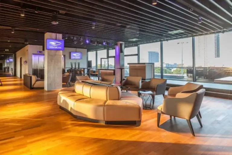 Eine moderne Lounge mit beigen Ledersofas und -sesseln auf einem Holzboden, mit großen Fenstern mit Blick auf die Stadt und an der Wand befestigten Bildschirmen mit blauem Logo. Der geräumige, stilvolle Bereich ist von natürlichem Licht durchflutet.