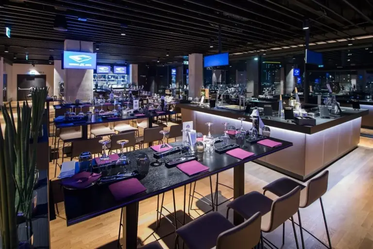 Ein modernes Restaurantinterieur mit eleganter Beleuchtung, schwarzen Tischen mit violetten Servietten und gepolsterten Stühlen. Der Raum verfügt über große Fenster, einen sichtbaren Barbereich und ein schlichtes, modernes Design.