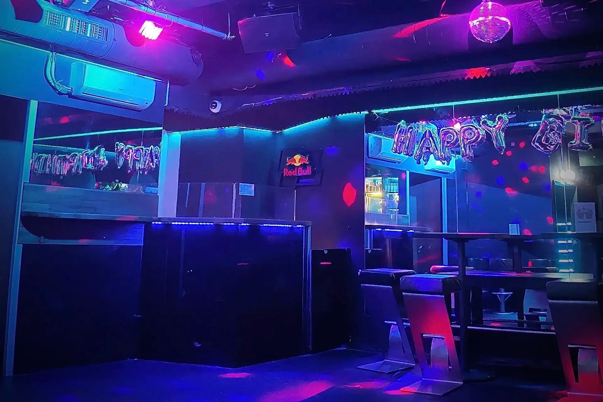 Gimme Five Ein schwach beleuchteter Nachtclub mit neonblauen und rosafarbenen Lichtern, leeren Tischen und Stühlen, einer Discokugel und einem Happy-Birthday-Banner am Spiegel, die eine festliche und doch ruhige Atmosphäre schaffen.