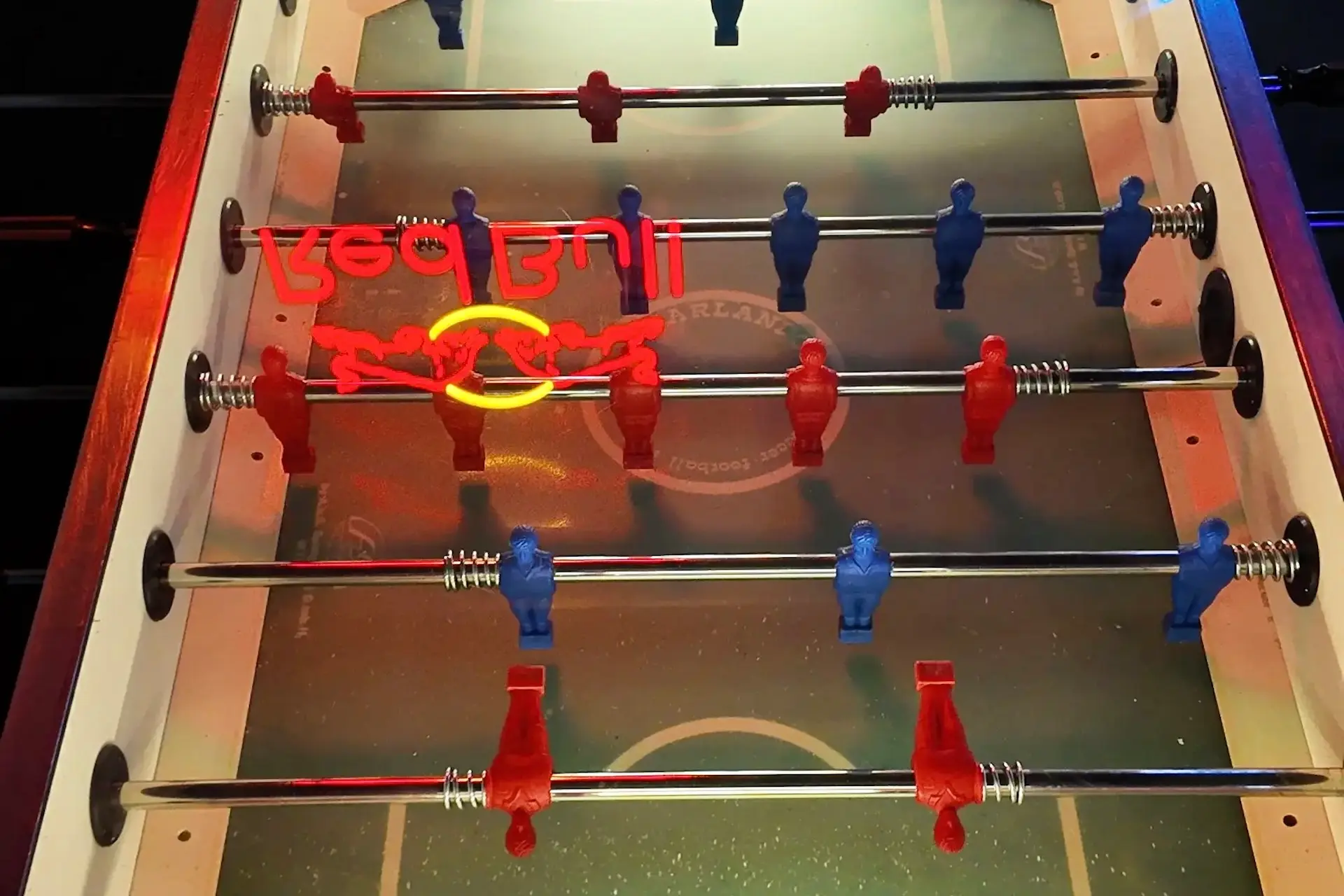 Ein Kickertisch mit roten und blauen Spielerfiguren unter heller Beleuchtung. Der Schriftzug Red Bull und ein Stierlogo spiegeln sich in Rot und Gelb auf der gläsernen Spielfläche.