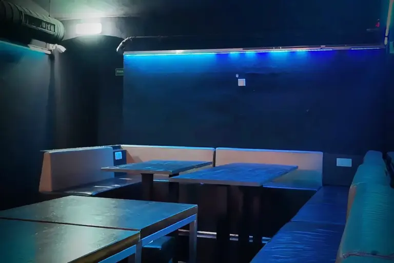 Eine schwach beleuchtete Lounge mit leeren Tischen und gepolsterten Sitzbänken an den Wänden. Blaue und weiße Lichter erhellen den dunklen Raum und schaffen eine ruhige und stille Atmosphäre.