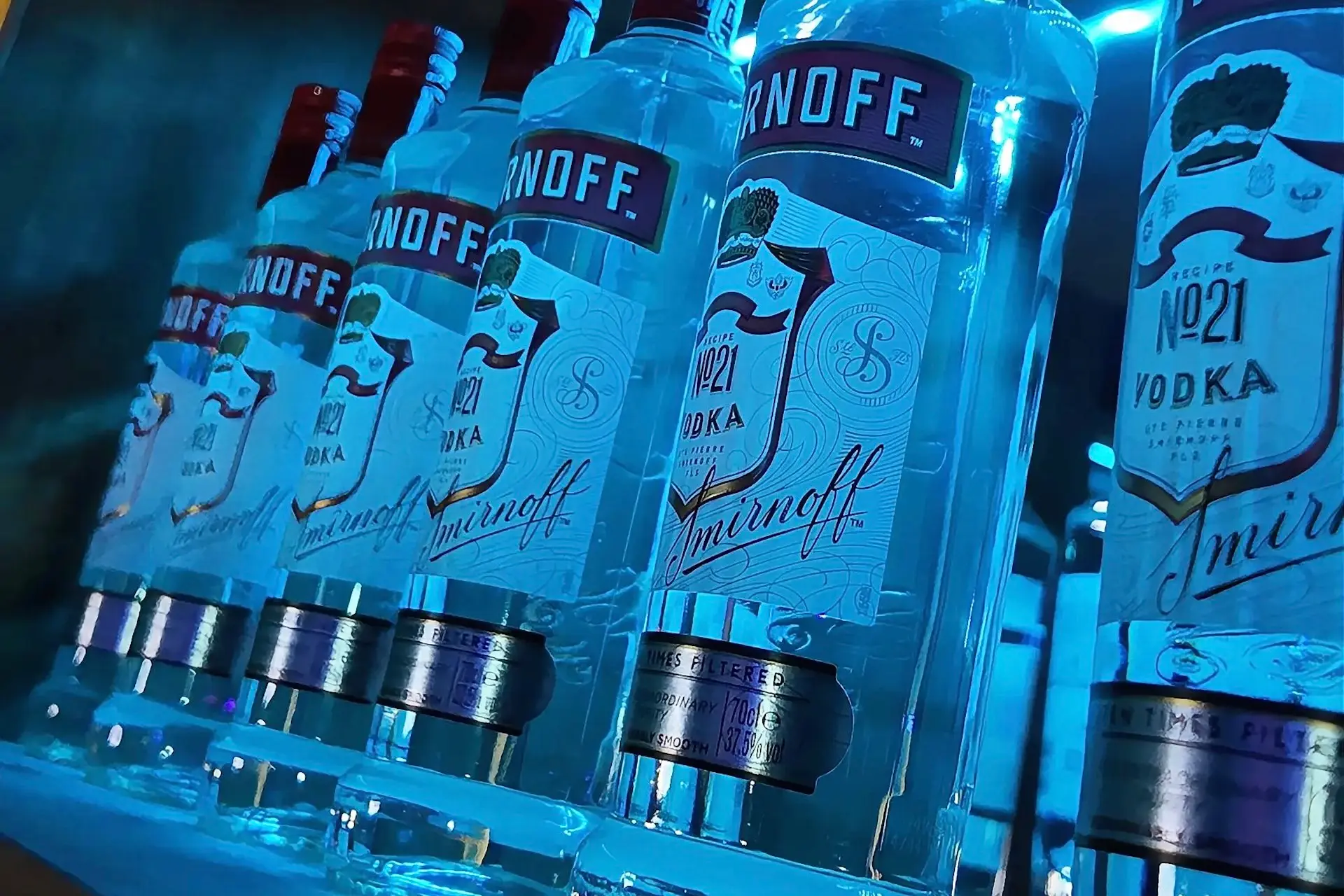 Mehrere Flaschen Smirnoff No.21 Wodka stehen in einer Reihe auf einem Regal und werden von blauem Licht angestrahlt. Die Etiketten und roten Flaschenverschlüsse sind deutlich sichtbar.