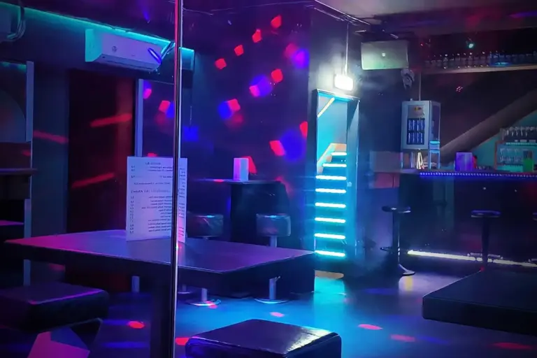 Leeres Nachtclub-Interieur mit bunten Neon- und LED-Lichtern, beleuchtetem Barbereich, Barhockern, Tischen und Ständen, moderner Einrichtung und einer Speisekarte auf einem der Tische. Es sind keine Menschen anwesend.