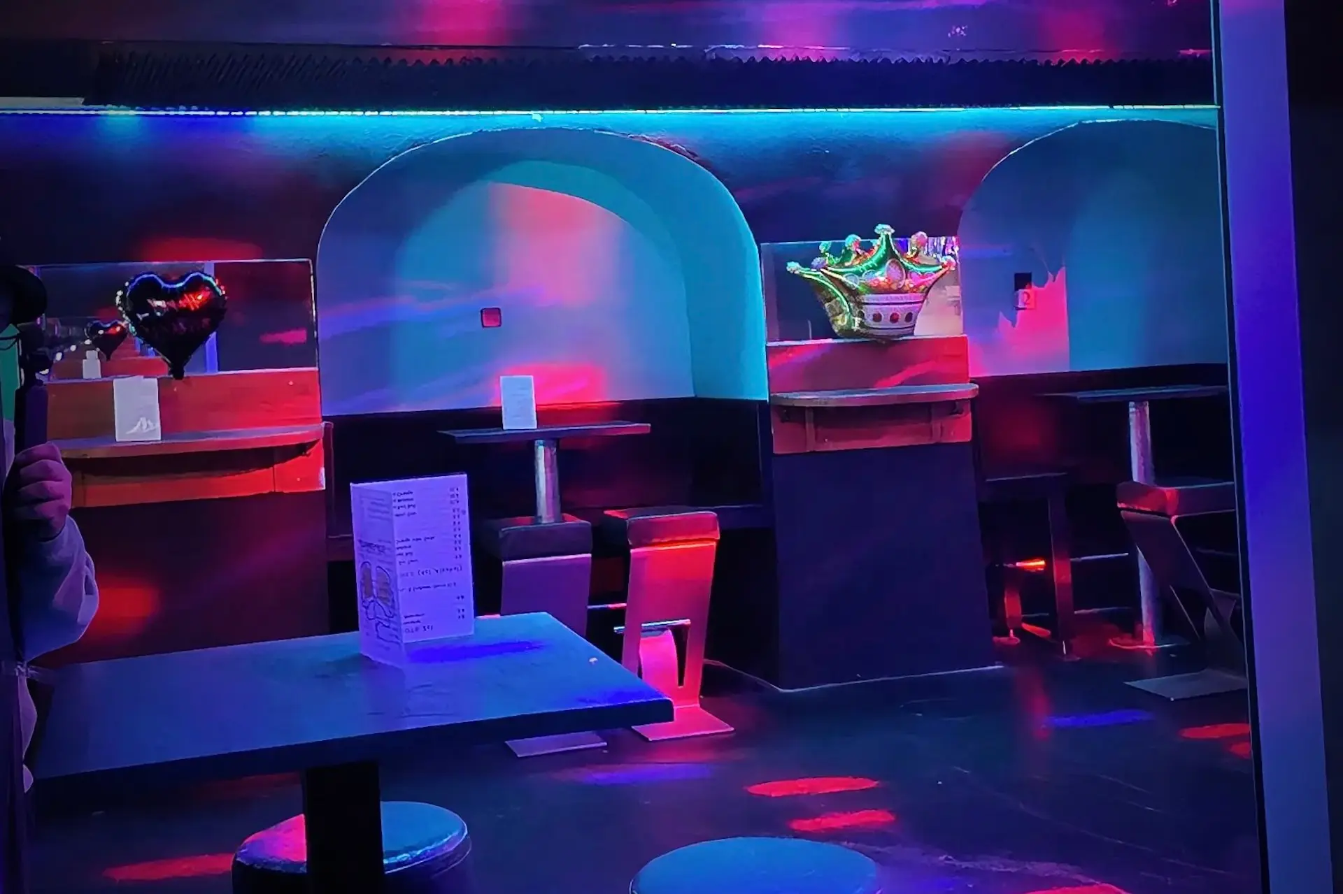 Das Interieur eines Nachtclubs mit blauer und pinker Neonbeleuchtung, leeren Ständen mit hohen Tischen und Hockern, einer Kronendekoration an einem Stand und Speisekarten auf den Tischen schafft eine lebendige, farbenfrohe Atmosphäre.
