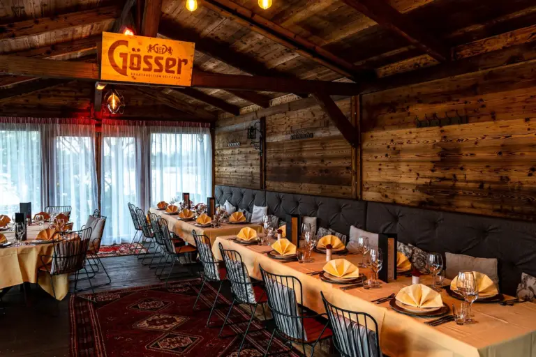 Gemütliches Holzrestaurant mit langen Tischen, die zum Essen gedeckt sind, mit ordentlich gefalteten Servietten, Glaswaren und Drahtstühlen. Die warme Beleuchtung und die rustikale Einrichtung schaffen eine einladende Atmosphäre. Ein Gösser-Bierschild hängt von der Decke.