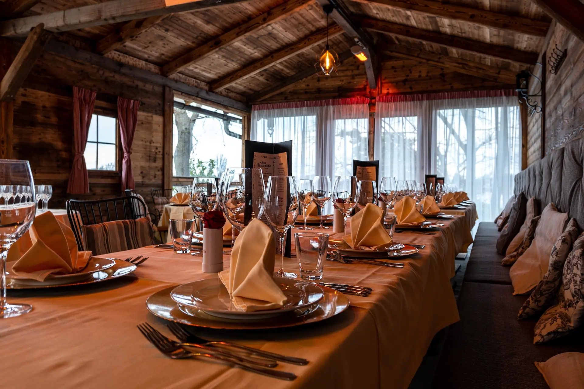 Elegantes Esszimmer mit Holzdecke und großen Fenstern. Der lange Tisch ist mit Tellern, Servietten, Gläsern und Silberbesteck gedeckt, bereit für ein formelles Essen. Die warme Beleuchtung schafft eine gemütliche Atmosphäre.