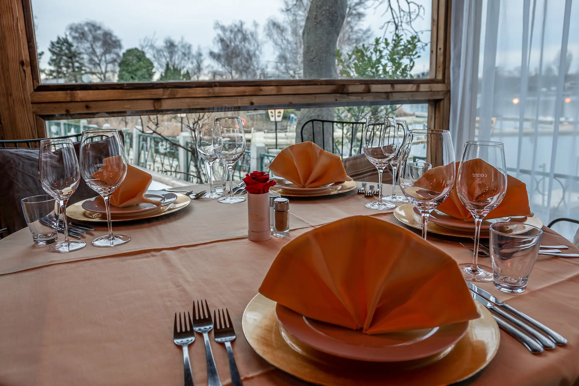 Ein für vier Personen gedeckter Esstisch mit orangefarbenen Servietten, die elegant auf Tellern gefaltet sind, umgeben von Besteck, Gläsern und einem kleinen roten Blumenschmuck, mit Blick ins Freie durch große Fenster.