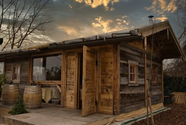 Eine rustikale Holzhütte mit offener Tür und kleinen Fenstern steht auf einer hölzernen Veranda, flankiert von Fässern, während die Sonne hinter ihr untergeht und ein warmes Licht auf die verstreuten Wolken am Himmel wirft.